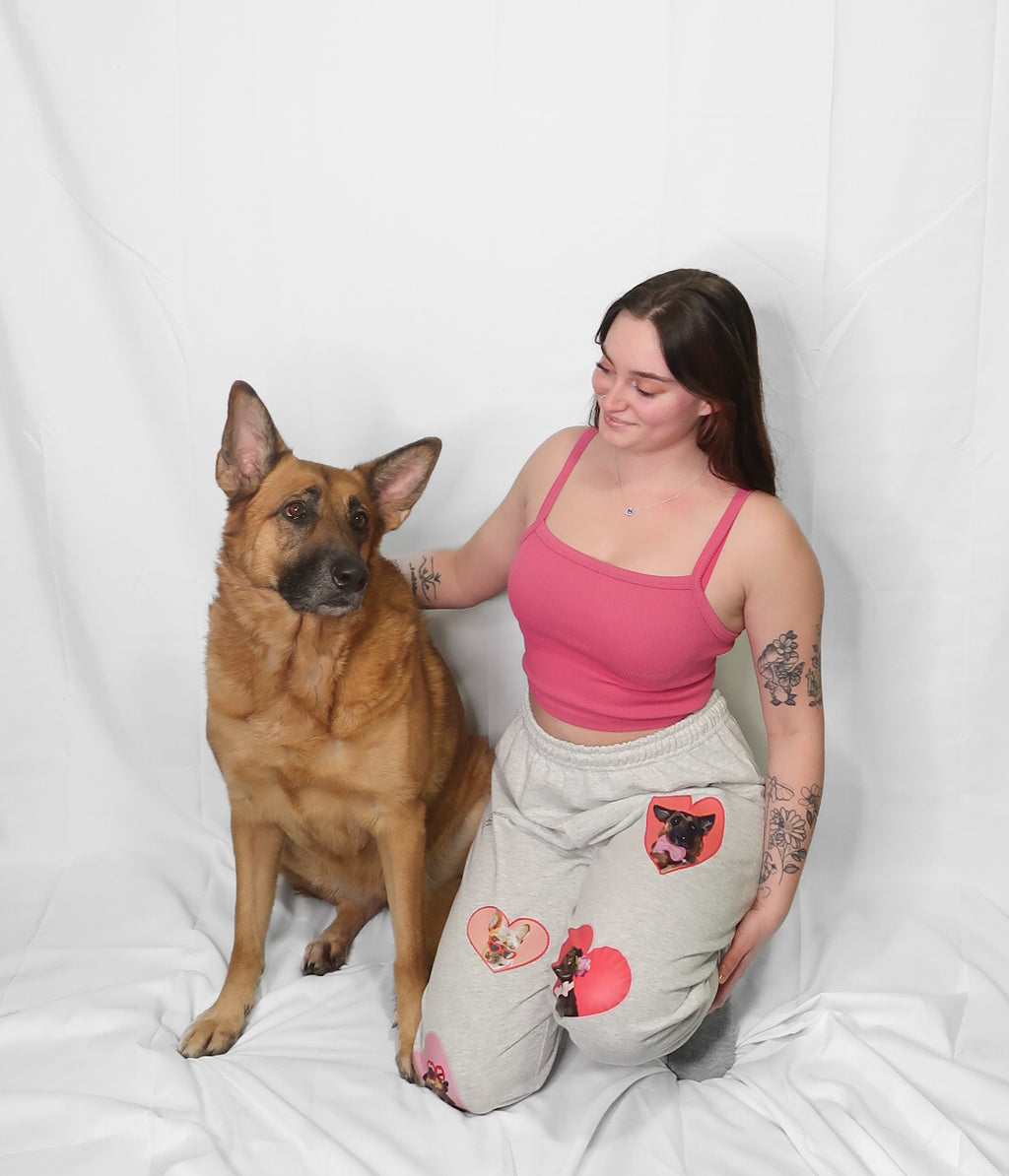 Woman in pink top and heart-patterned pants sitting next to a German Shepherd on a white background